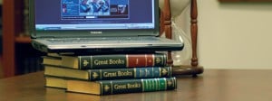stack of books, labeled Great Books, atop a table with a laptop resting on top of them, and an hourglass in the background