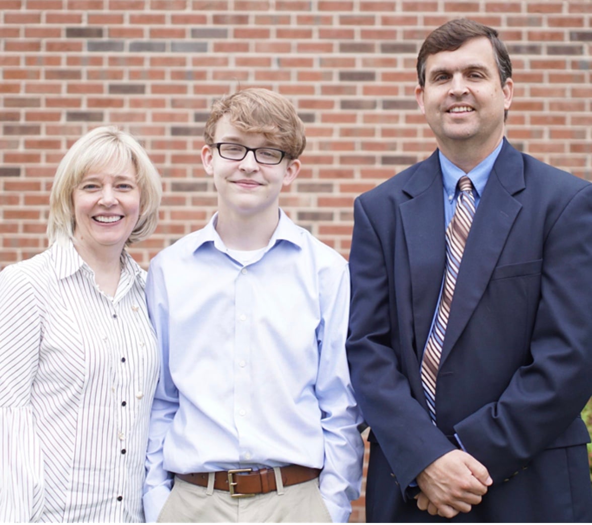 Blonde boy in light dress shirt standing in between mother in pink shirt and father in a suit.