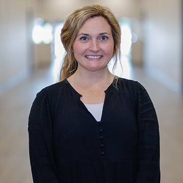 Woman with blonde hair pulled into ponytail smiling into camera while wearing black long sleeved shirt.