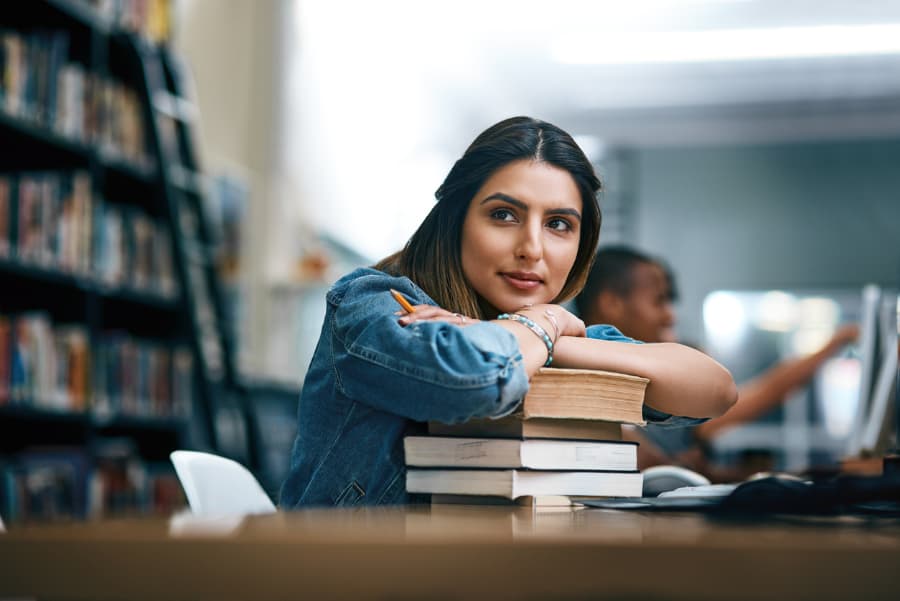 A student rests her chin on her arms atop a stack of books in a library