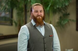 A man with red hair and a beard is wearing a light blue dress shirt and a gray vest while smiling.