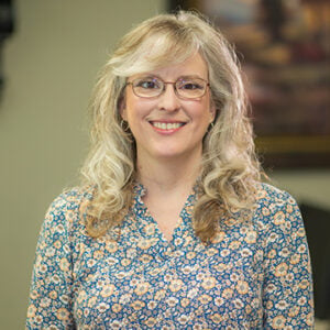A woman with silver hair and red highlights is wearing glasses and a blue and yellow floral shirt while smiling into the camera.