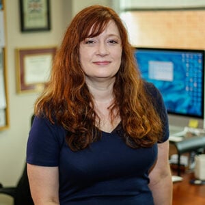 Woman with long brown hair smiling into camera wearing a navy blue shirt.
