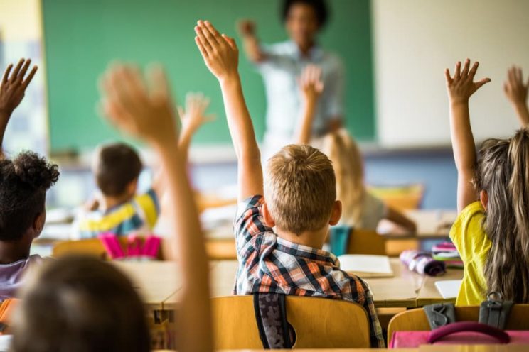A room of students with raised hands while teacher selects one. 