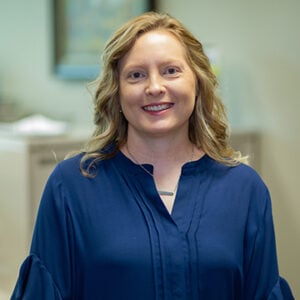 A woman with long blonde hair wearing a silver necklace and a navy blue long sleeved top is smiling.