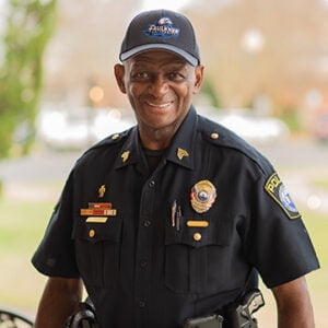 A man is standing outside wearing a Faulkner hat and a sergeant's uniform while smiling at the camera.