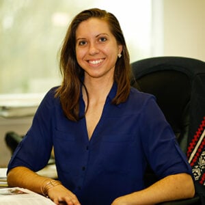 A woman with long brunette hair is wearing a navy blue shirt and smiling.