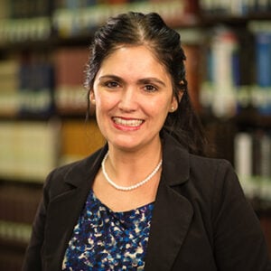 A woman with long dark hair pulled back in ponytail is wearing a silver necklace, a blue shirt, and a black blazer while smiling.