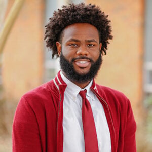 A man with short black hair and a beard is wearing a white dress shirt with a red tie and sweater while smiling.