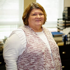 A woman with short blonde hair wearing a white long sleeved shirt with a light pink cardigan while smiling.
