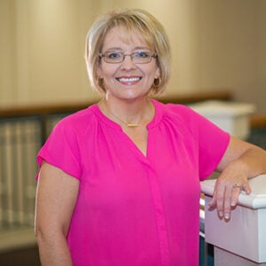 A woman with short blonde hair is wearing glasses, a gold pendant, and a hot pink shirt while smiling.