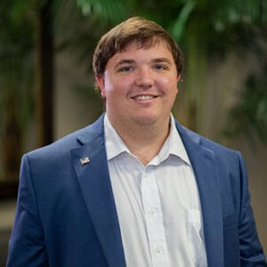 A man with short brown hair is smiling at the camera while wearing a white dress shirt and a navy blue blazer.
