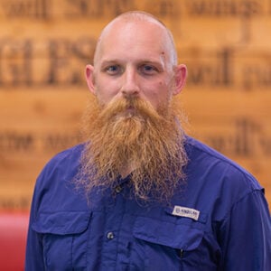 A man with a long read beard is wearing a royal blue button up shirt while smiling at the camera.