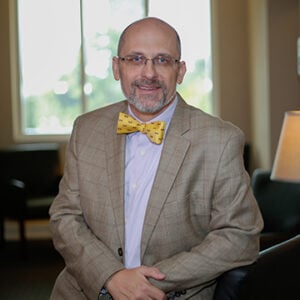 A man with a salt and pepper beard is wearing glasses, a light purple dress shirt, a light brown blazer, and a yellow bowtie while smiling at the camera.