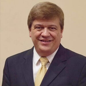 A man with short brunette hair is wearing a white dress shirt, a black blazer, and a yellow tie while smiling at the camera.