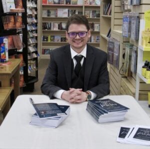 A man with short brown hair and glasses is wearing a black suit with a black tie smiling while sitting at a table covered in books.