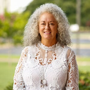 Woman with long curly silver hair is wearing a white lace top and a long silver necklace while smiling into the camera.