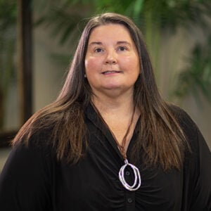 A woman with long brunette hair is wearing a silver necklace and a black dress while smiling at the camera.
