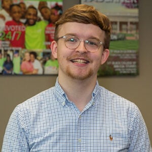 A man with short dirty blonde hair and glasses is wearing a blue and white checkered dress shirt and smiling at the camera.