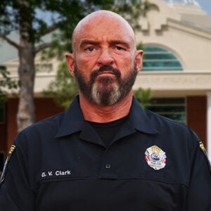 A man with a salt and pepper beard is looking at camera while wearing a black shirt with his name and badge.