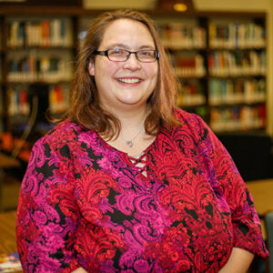 A woman with shoulder length brunette hair is wearing glasses and a black, red, and pink floral shirt while smiling at the camera.