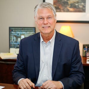 A man with silver hair is wearing a light blue dress shirt and a navy blue blazer while smiling into the camera.