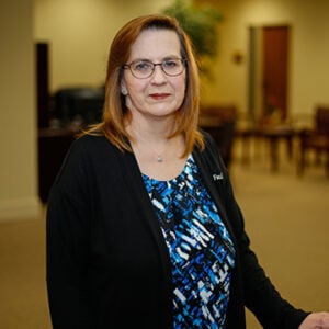 A woman with shoulder length red hair wearing glasses, a blue and black swirl patterned shirt, and a black blazer smiling into the camera.