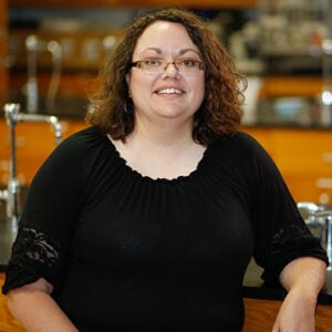 A woman with shoulder length curly brunette hair is wearing glasses and a black shirt while smiling at the camera.
