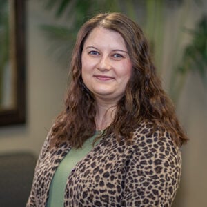 A woman with long brunette hair is wearing a green shirt and a cheetah print cardigan while smiling.