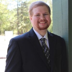 A man with short red hair and beard is wearing a black suit while smiling.