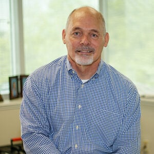 A man with a salt and pepper goatee is wearing a white and blue checkered dress shirt while smiling at the camera.