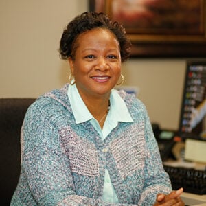 A woman with short brunette hair is wearing a light green dress shirt with a light blue cardigan while smiling.