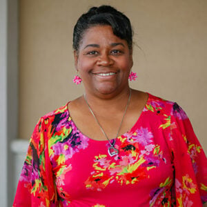 A woman with short black hair is wearing pink earrings, a silver necklace, and a multi coloured shirt while smiling.