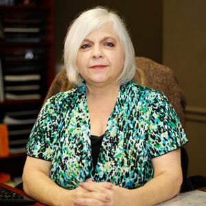 A woman with short white hair is wearing a green multi-coloured top while smiling.