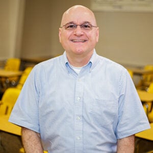 A man wearing glasses and a light blue dress shirt is smiling.