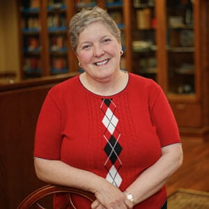 A woman with short silver hair is wearing a red sweater with white and black diamonds while smiling.