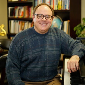 A man with short brunette hair is wearing glasses and a blue sweater while smiling.