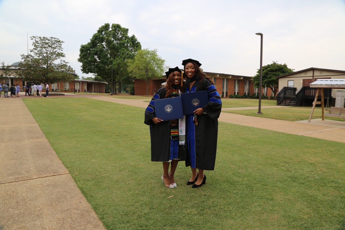 l-r Katie Jackson and Alysia (Perkins) Cutts stand on the grassy area outside Faulkner's gym in their graduation robes.