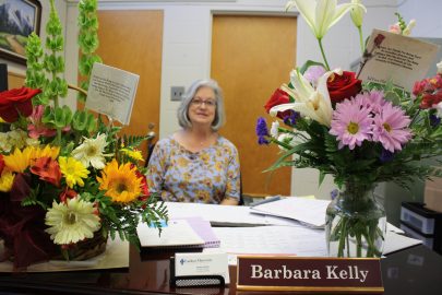 Barbara Kelly sits at her desk surrounded by two large bouquet of flowers.