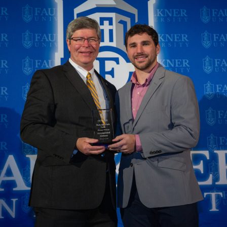 President Mitch Henry presents Hunter Wilson with the Accomplished Alumnus Award in front of a Faulkner blue backdrop.