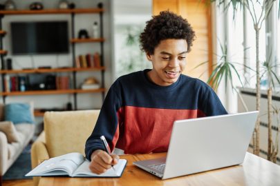 Student smiles while engaging with laptop computer screen and taking notes by hand at home