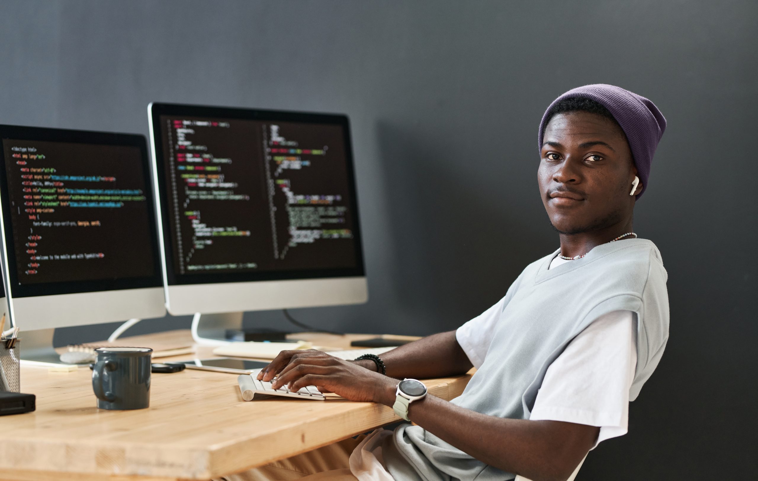 Young black man in casualwear keeping hands on computer keyboard while sitting in front of monitors and looking at camera